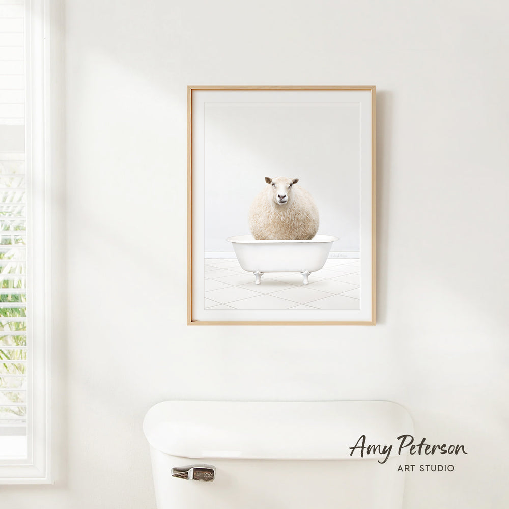 A framed image of a sheep sitting in a bathtub is hanging on a white wall above a white toilet.