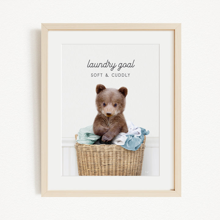 A cute brown bear sitting in a wicker laundry basket filled with clothes, with the text "laundry goal SOFT & Cuddly" displayed above the bear.