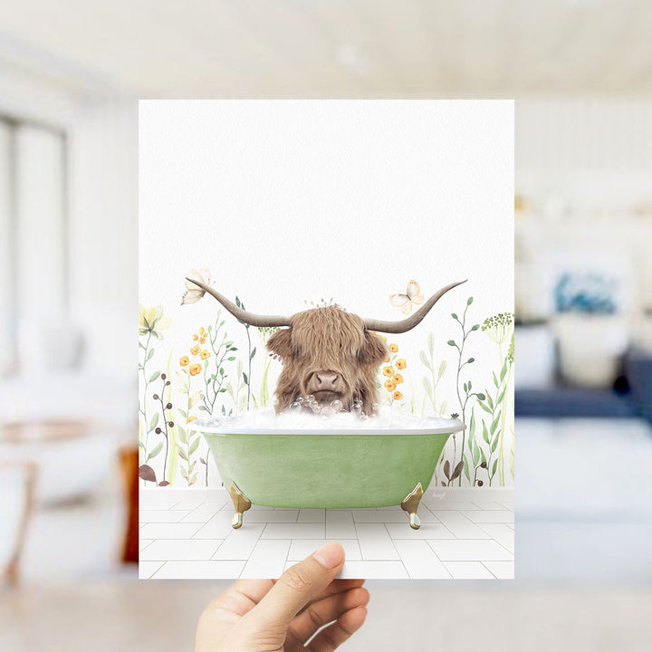 A hand holds up a framed image depicting a Highland cow with large horns sitting in a green bathtub filled with bubbles, surrounded by a floral background.