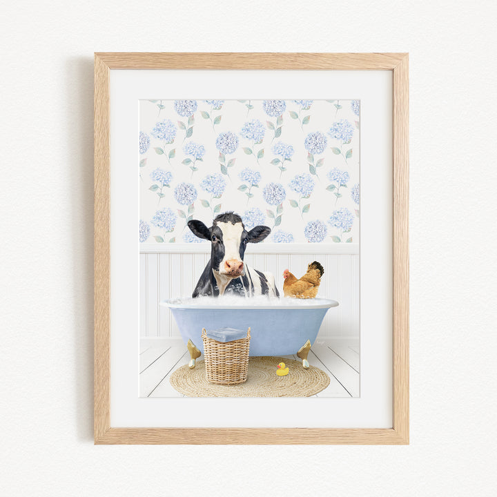 A framed image depicting a cow sitting in a bathtub, surrounded by a chicken and a duck, with a blue floral patterned wall in the background.