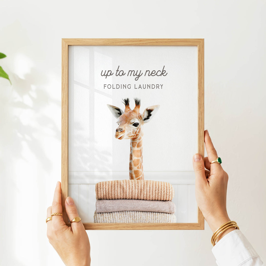 A person's hands holding a framed picture of a giraffe with folded clothes underneath it, against a white background.