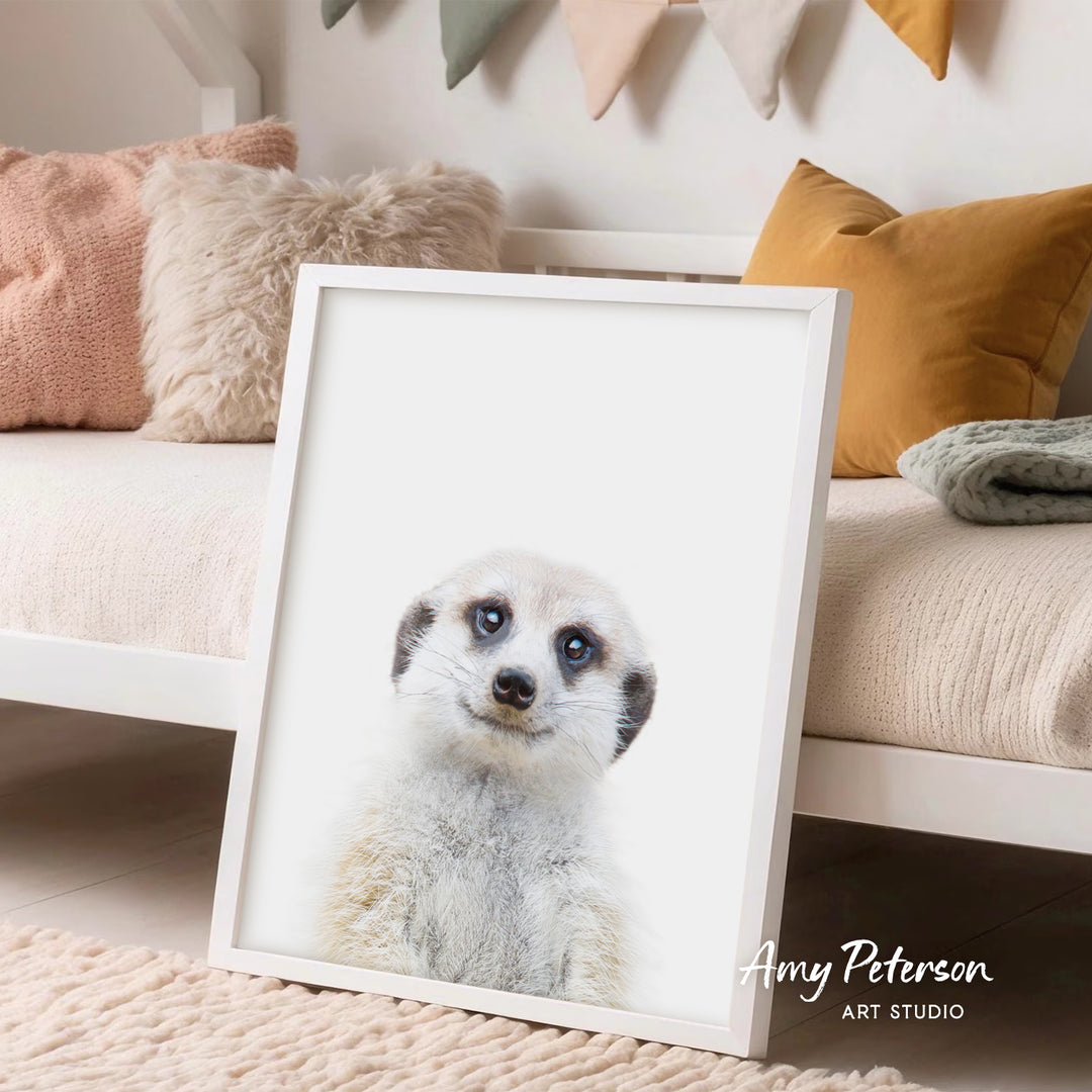 A framed print of a meerkat's face is displayed in a room with a white bed and colorful pillows.