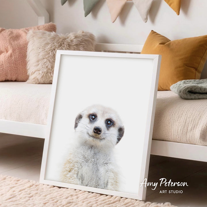 A framed print of a meerkat's face is displayed in a room with a white bed and colorful pillows.