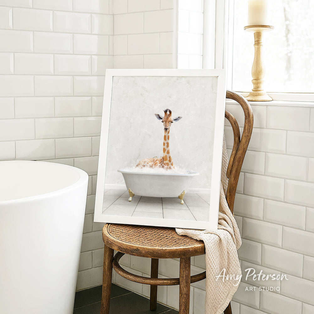 A framed image of a giraffe sitting in a bathtub is displayed on a wooden chair in a bathroom setting.