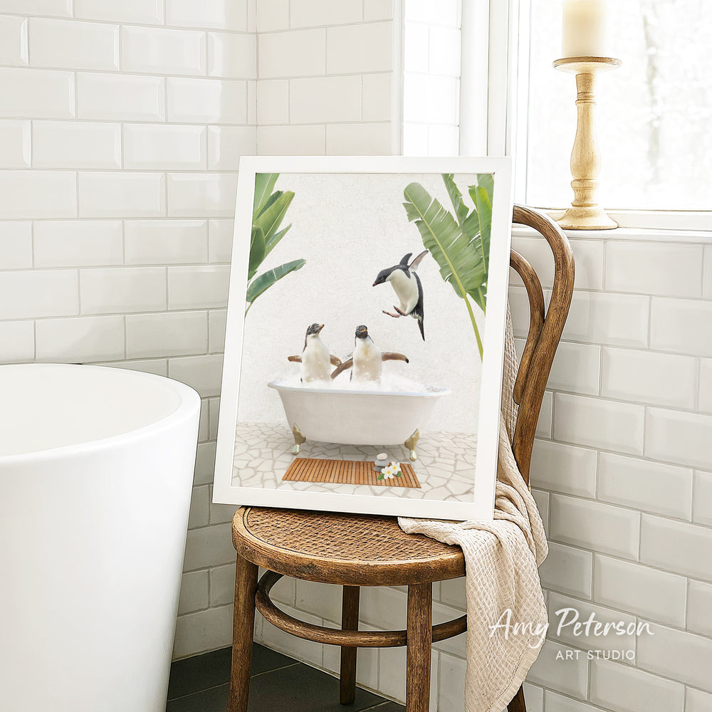 A framed image of two penguins sitting in a bathtub, surrounded by tropical plants, is displayed on a wooden chair in a bathroom setting.
