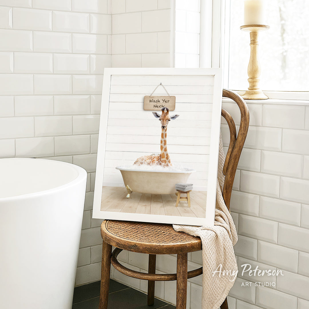 A framed image of a giraffe sitting in a bathtub, with a sign that says "Wash Your Neck" hanging above it. The image is displayed on a wooden chair in a bathroom setting.