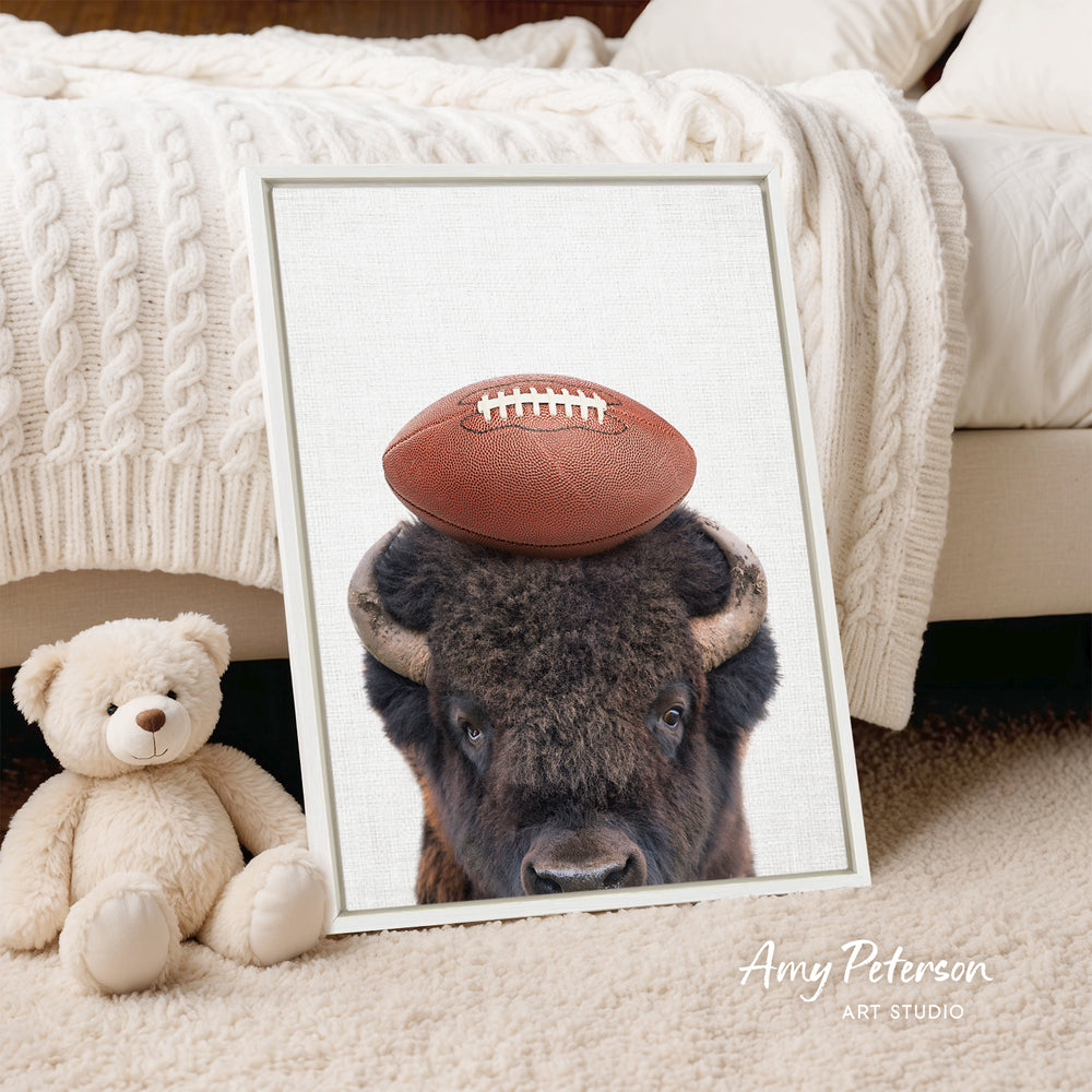 A framed print of a buffalo resting its head on a football is displayed on a bed, with a teddy bear next to it.