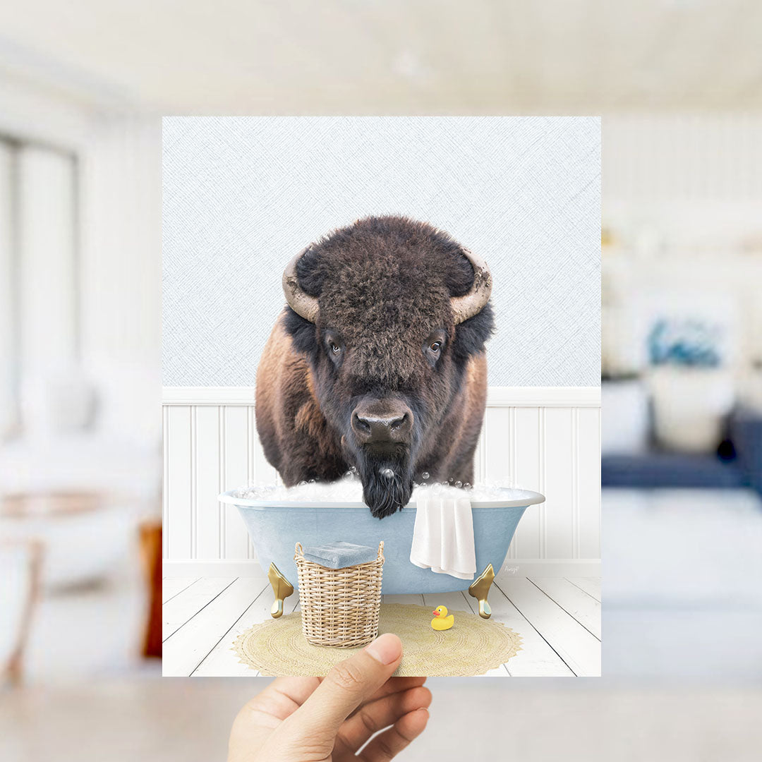 A buffalo with large horns is sitting in a bathtub filled with water, surrounded by a yellow rubber duck and a wicker basket.