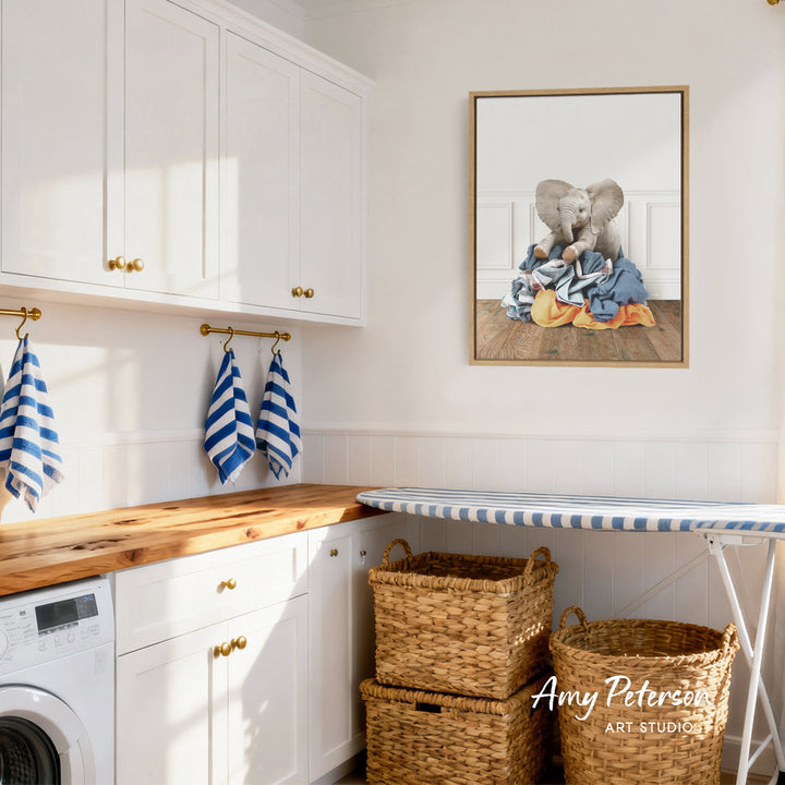 A bright and airy laundry room with white cabinets, a wooden countertop, and a framed picture of two elephants on a pile of clothes hanging on the wall.