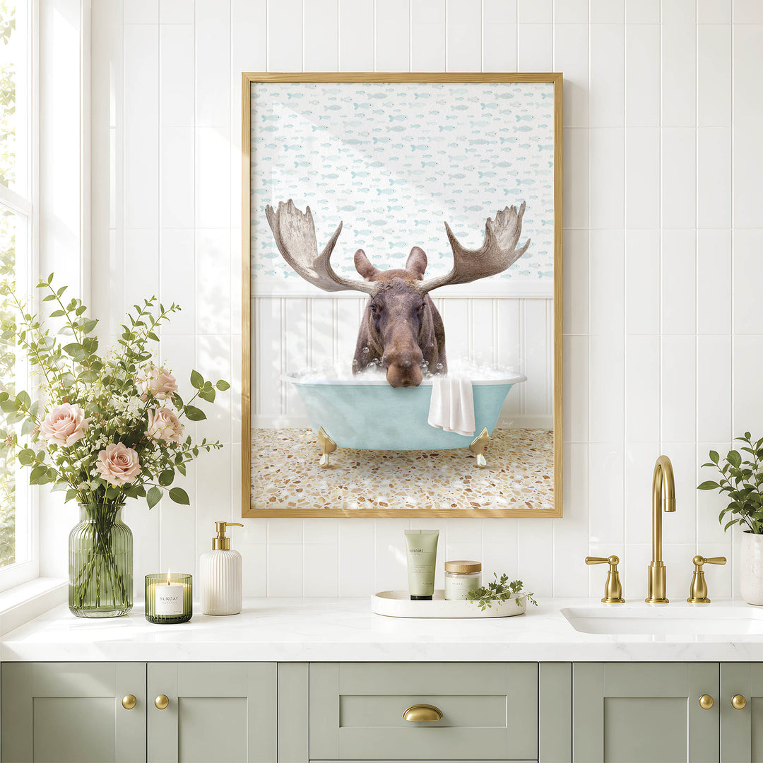 A framed image of a moose sitting in a bathtub, surrounded by a floral arrangement and various bathroom accessories, such as a soap dispenser and a candle.