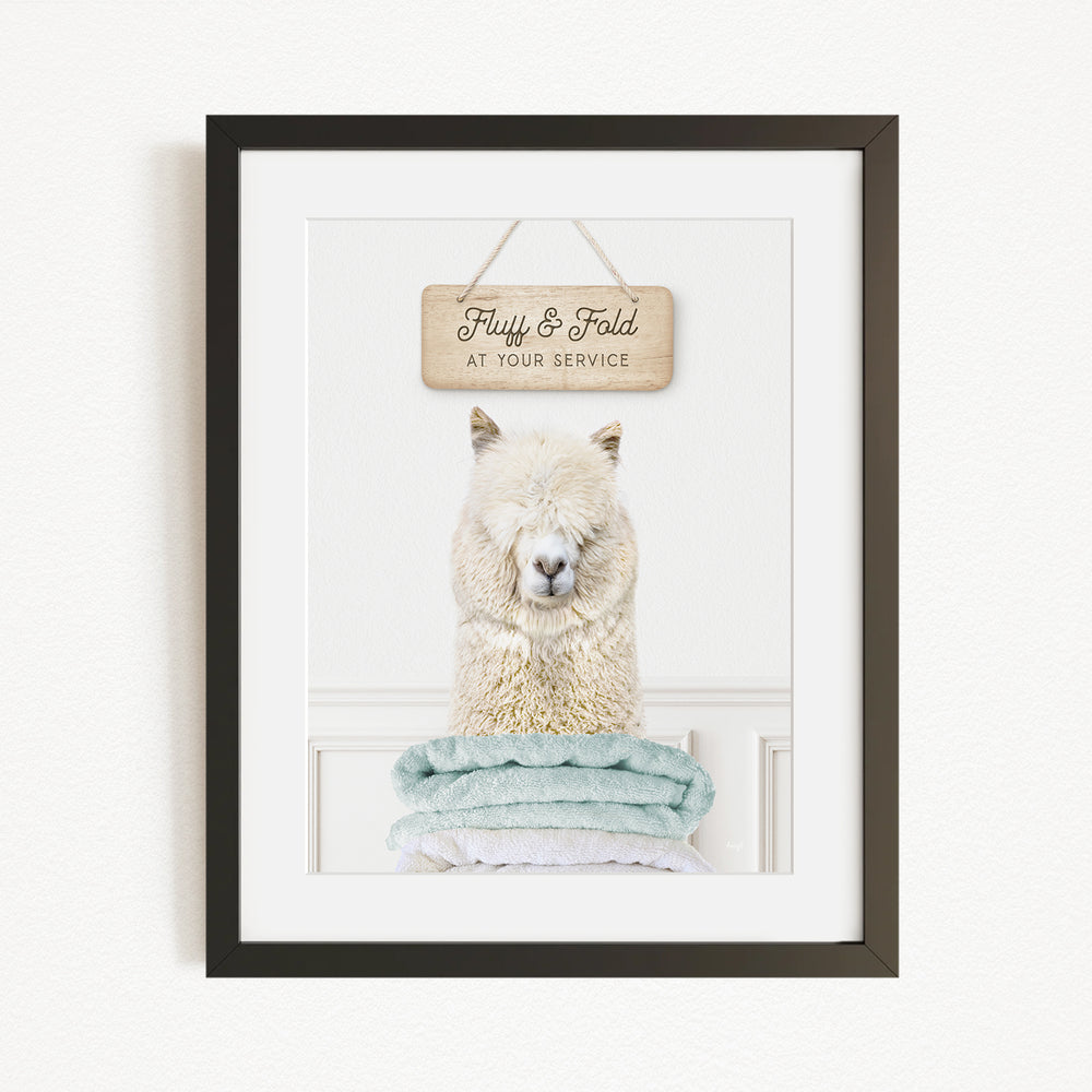 A framed image of a fluffy white alpaca sitting on a stack of blue towels, with a wooden sign above it that says "Fluffy & Fold at your service".