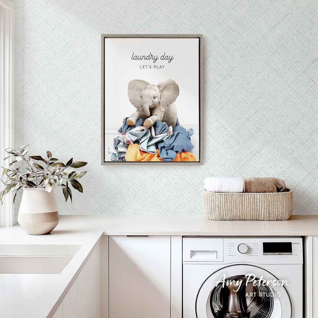 a framed poster of two elephants sitting on a pile of laundry, with the text "laundry day let's play" written above them. The poster is hanging on a wall above a white countertop with a potted plant and a basket of folded clothes.