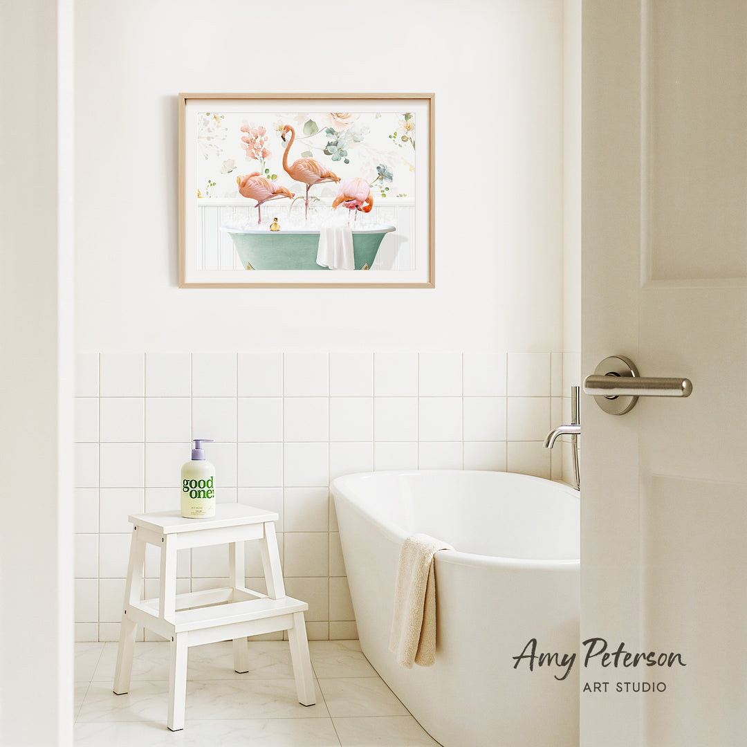 A framed painting of two pink flamingos in a green bathtub hangs on a white tiled wall above a white bathtub in a bathroom.