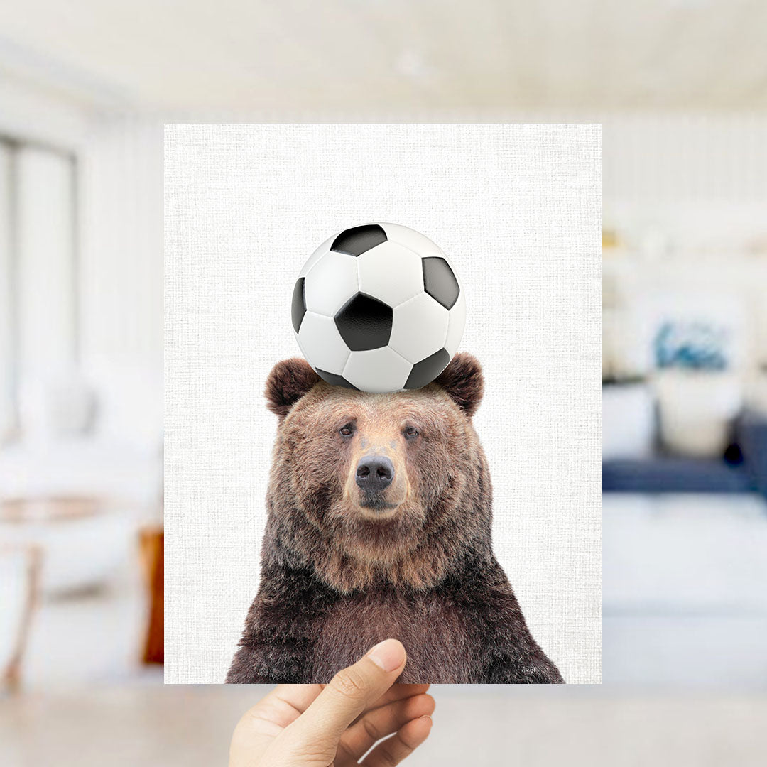 A hand holds up a picture of a brown bear with a soccer ball on its head.