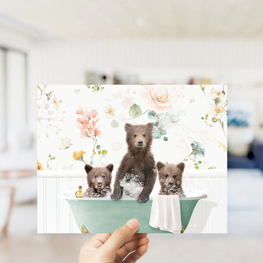 A hand holds up a framed image depicting three adorable brown bears sitting in a bathtub filled with bubbles, surrounded by a floral pattern.