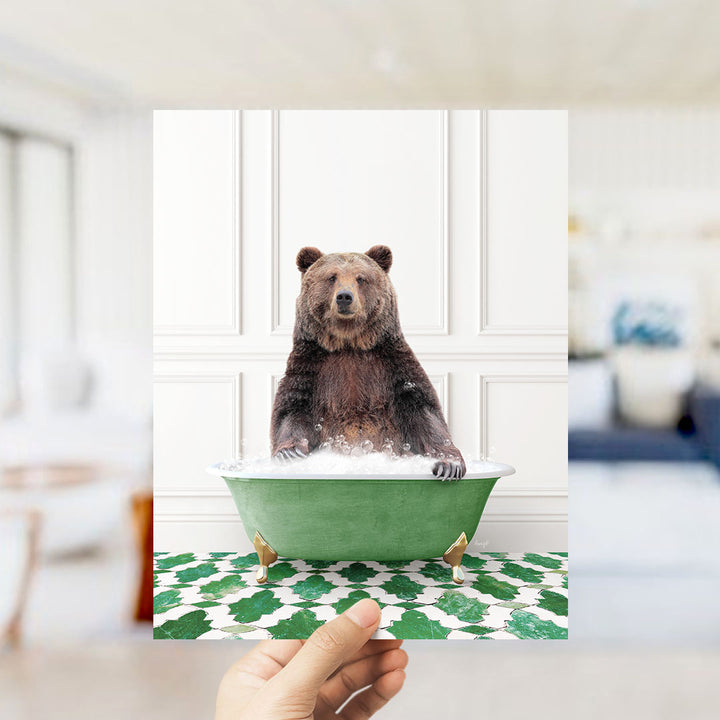 A hand holds up a framed image of a brown bear sitting in a green bathtub filled with bubbles, surrounded by a green and white patterned floor.