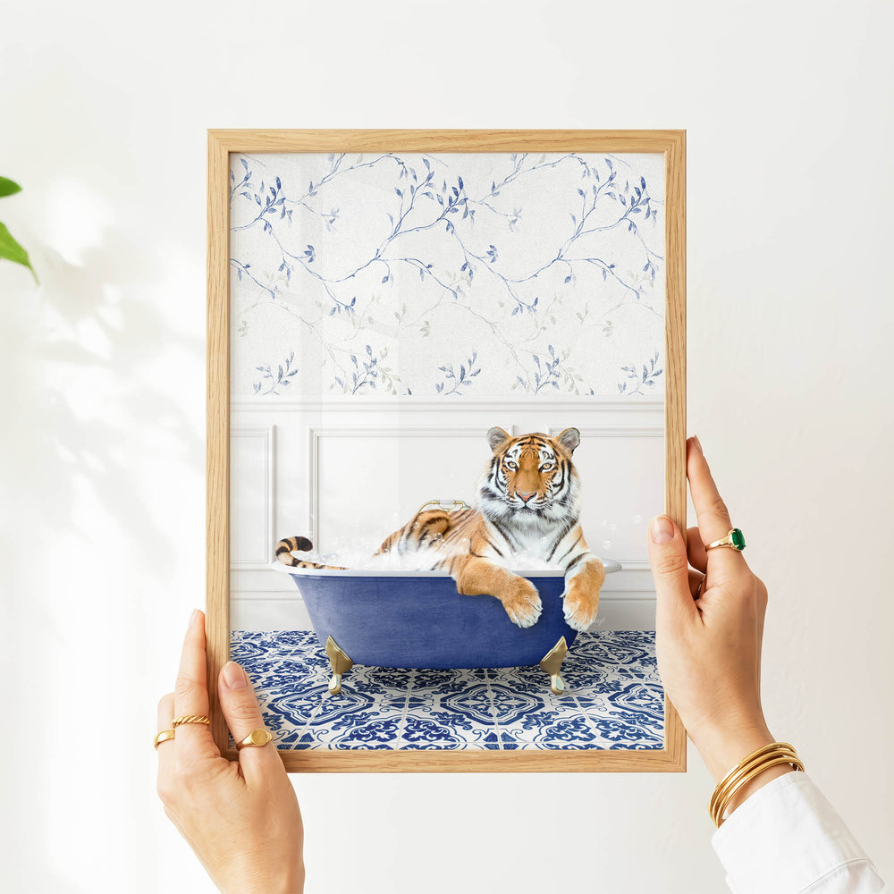 A person's hands are holding a framed picture of a tiger sitting in a blue bathtub, with a patterned wall behind it.