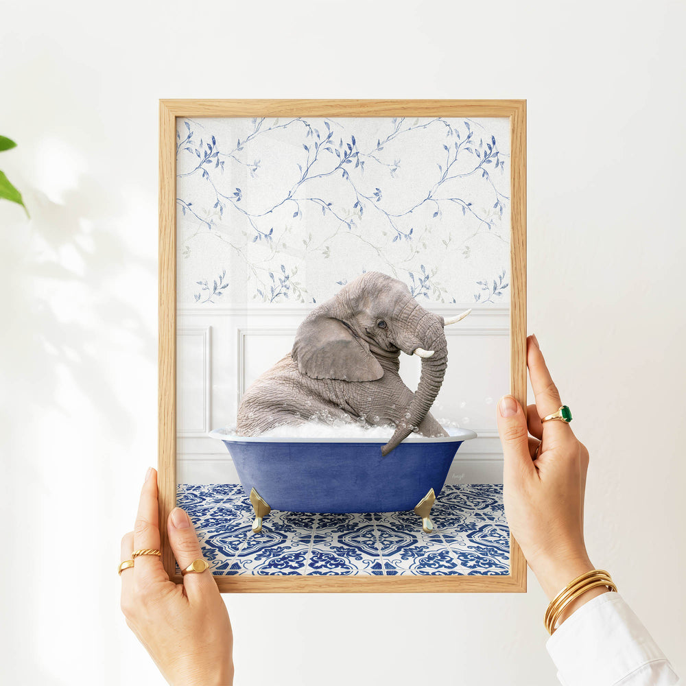 A person's hands are holding a framed picture of a gray elephant sitting in a blue bathtub, with a patterned wall behind it.