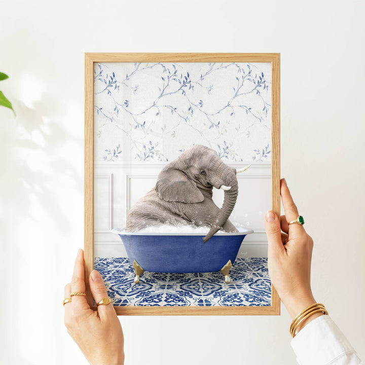 A person's hands are holding a framed picture of a gray elephant sitting in a blue bathtub, with a patterned wall behind it.