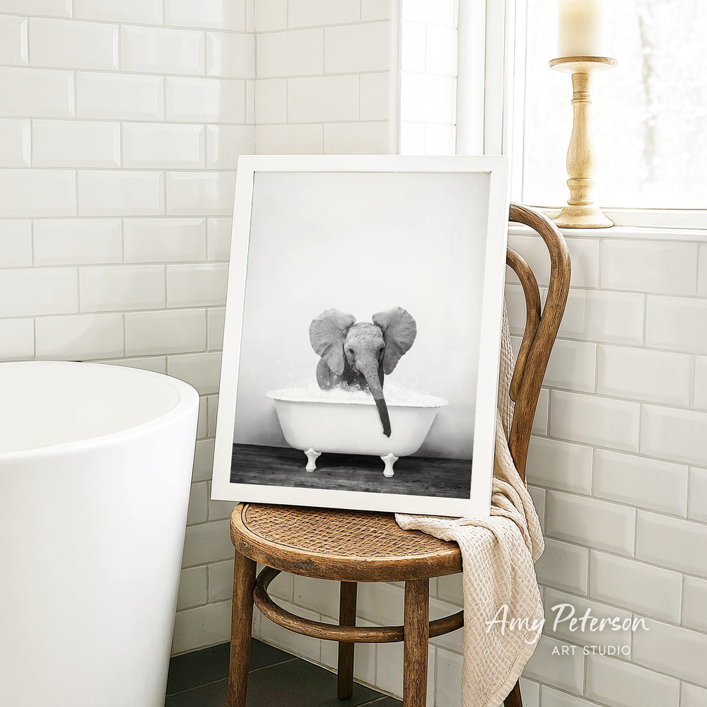 A framed black and white photograph of a baby elephant sitting in a bathtub, with a towel draped over the back of a wooden chair in a bathroom setting.