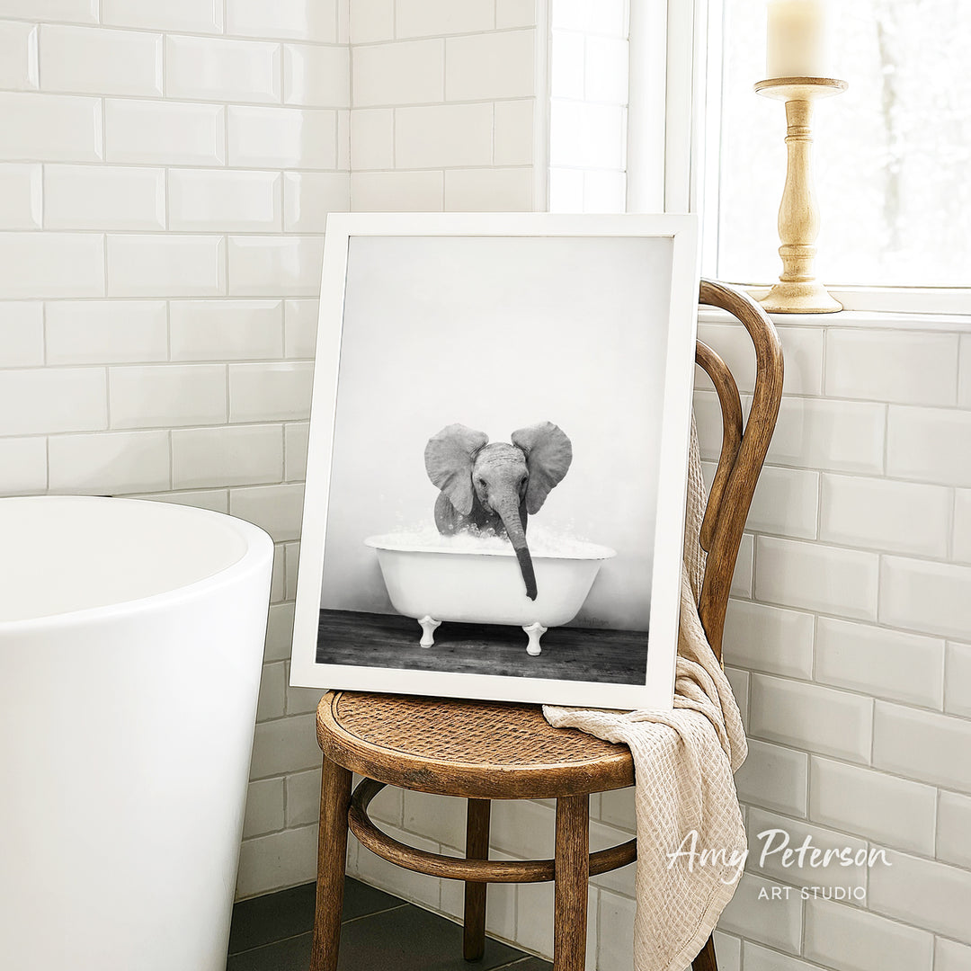 A framed black and white photograph of a baby elephant sitting in a bathtub, with a towel draped over the back of a wooden chair in a bathroom setting.