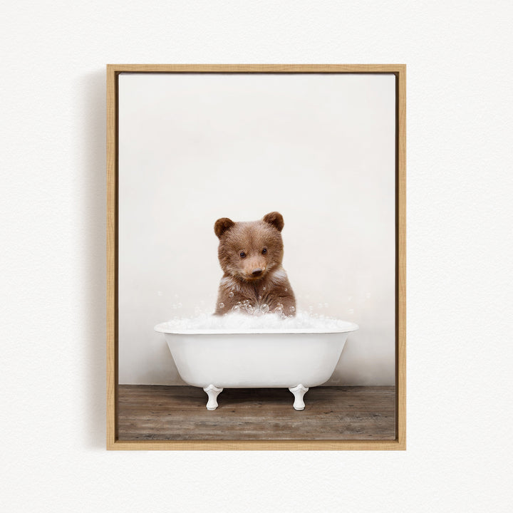 A cute brown bear cub sitting in a white bathtub filled with bubbles, looking directly at the camera.