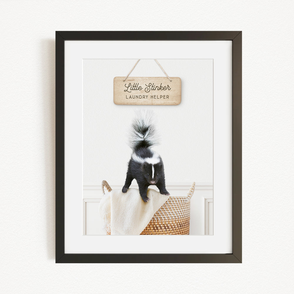 A framed photograph of a black and white skunk standing on a woven basket, with a wooden sign above it that reads "Little Slicker Laundry Helper".