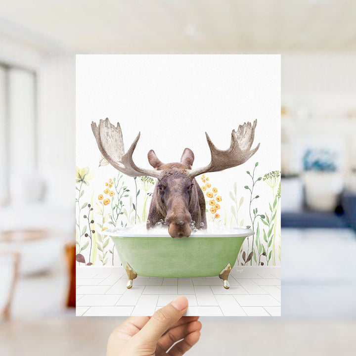 A moose with large antlers is sitting in a green bathtub filled with water, surrounded by a floral background.