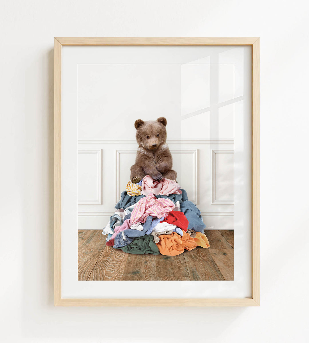 A brown teddy bear sits atop a pile of clothes on a wooden floor.