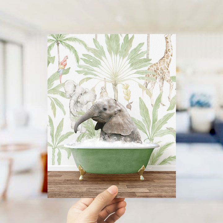 A hand holds up a framed image depicting a baby elephant sitting in a green bathtub, surrounded by tropical foliage and animals.