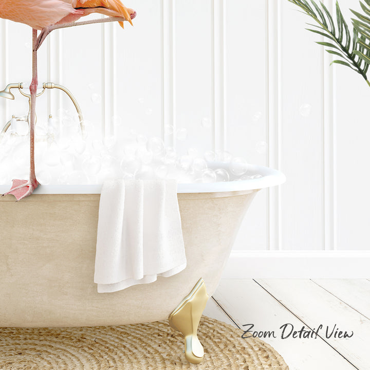 A white claw-foot bathtub filled with bubbles, with a towel hanging on the side and a pink flamingo perched on the edge of the tub.