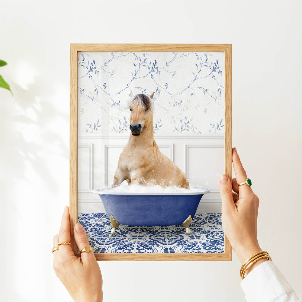 A person's hands are holding a framed picture of a horse sitting in a blue bathtub filled with water and bubbles.