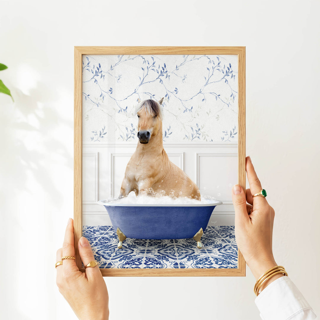 A person's hands are holding a framed picture of a horse sitting in a blue bathtub filled with water and bubbles.