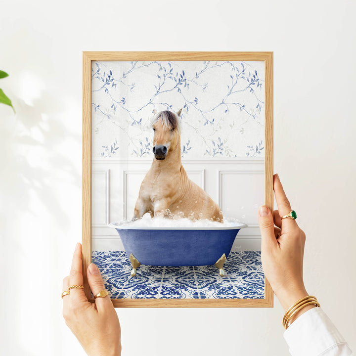 A person's hands are holding a framed picture of a horse sitting in a blue bathtub filled with water and bubbles.