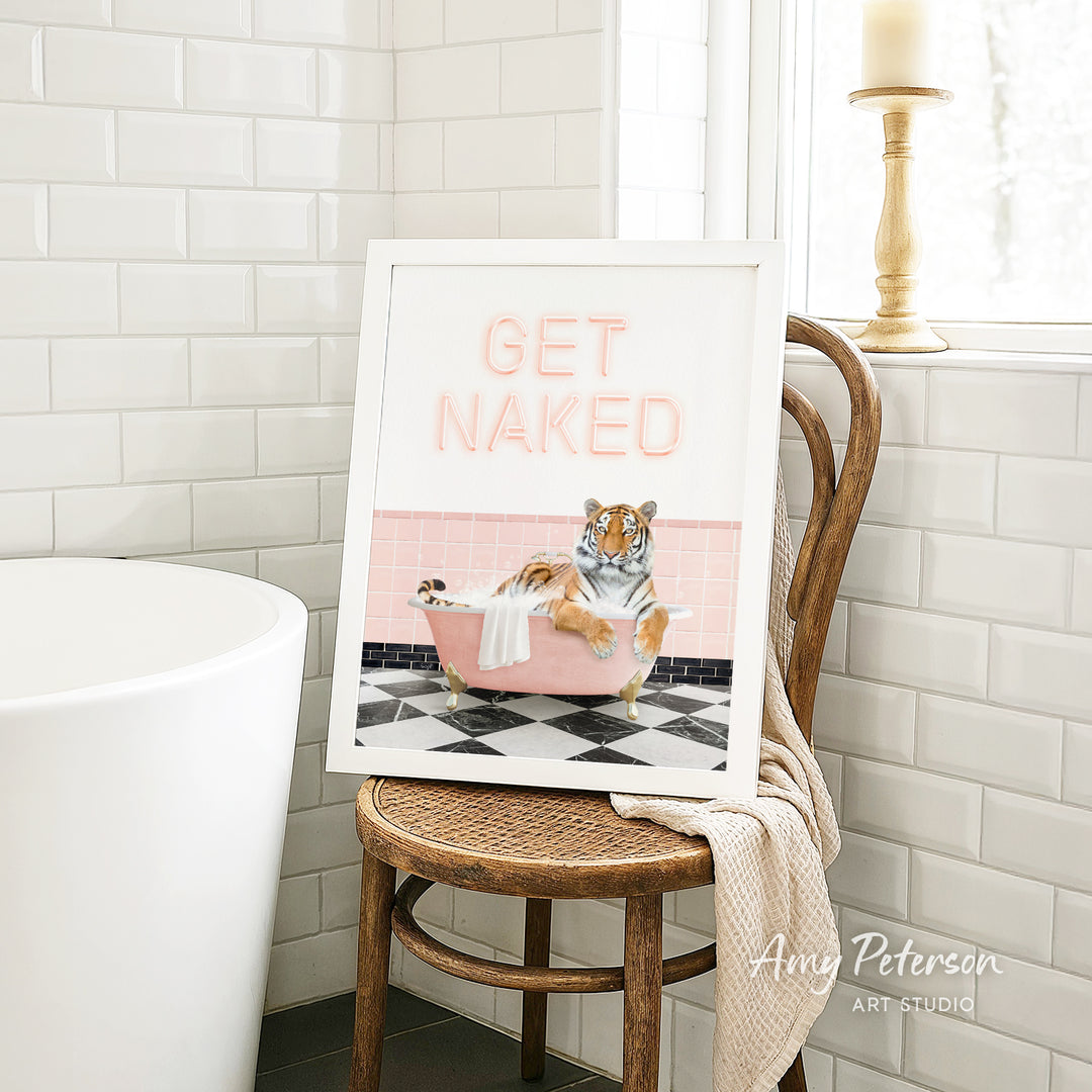 A framed print with a tiger sitting in a bathtub, surrounded by pink and black tiles, and the text "GET NAKED" is displayed on a wooden chair in a bathroom setting.