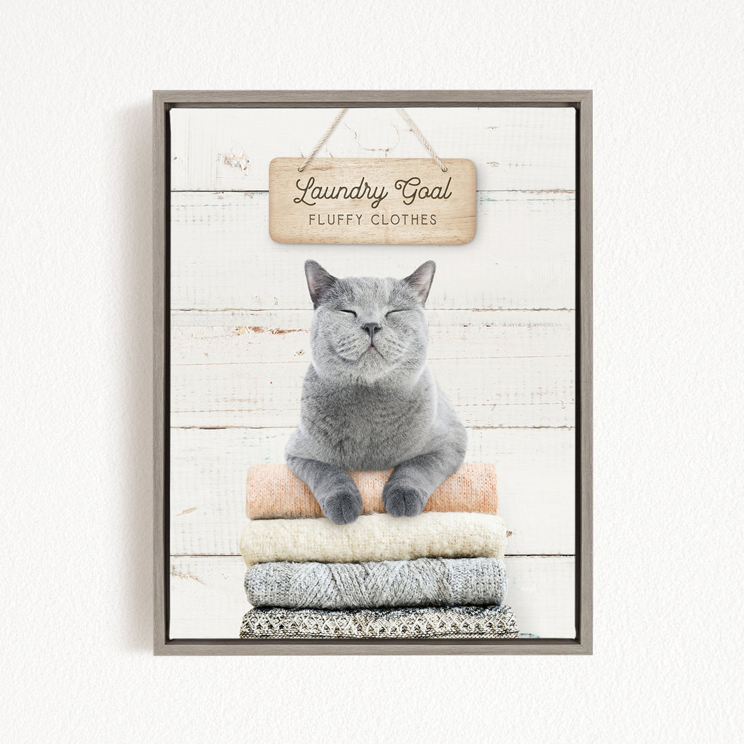 A gray cat sitting on a stack of laundry, with a wooden sign above that says "Laundry Goal Fluffy Clothes".