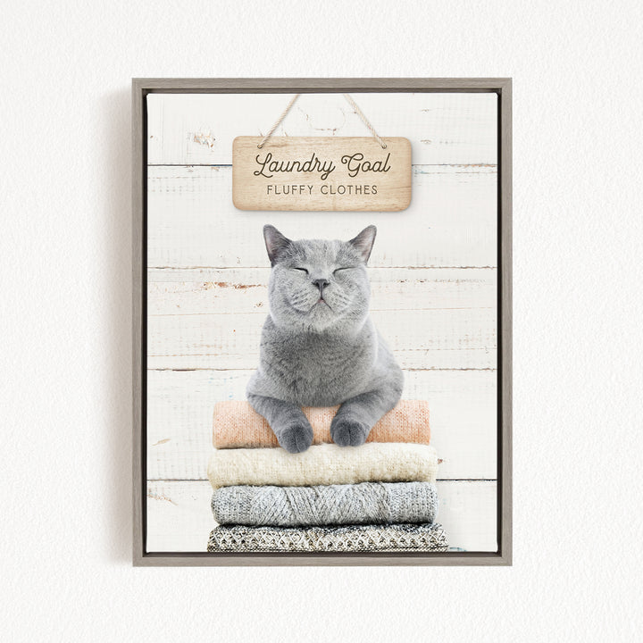 A gray cat sitting on a stack of laundry, with a wooden sign above that says "Laundry Goal Fluffy Clothes".