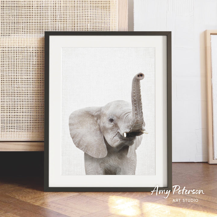 A framed photograph of a baby elephant with its trunk raised, displayed on a wooden floor against a white wall.