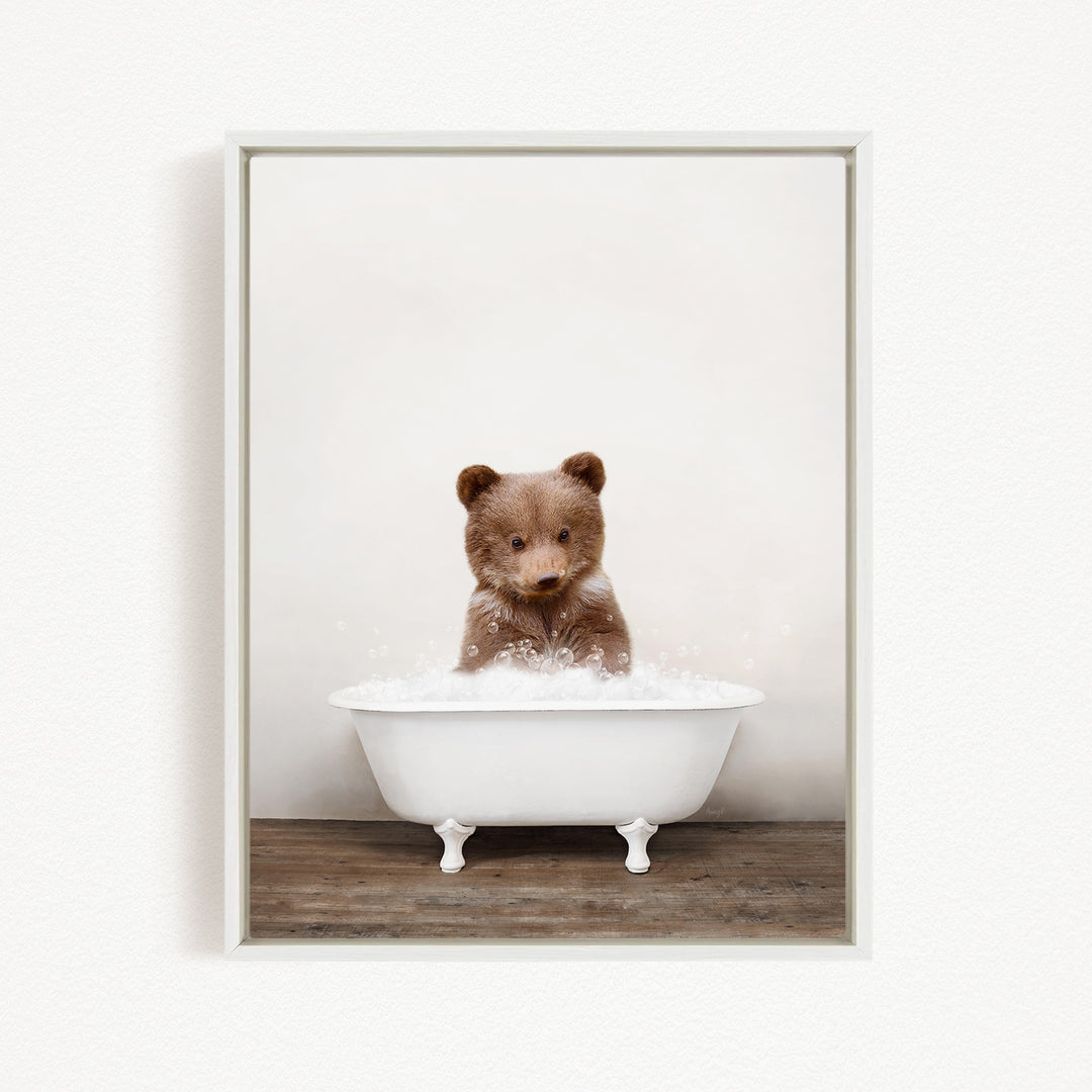 A cute brown bear sitting in a white bathtub filled with bubbles, looking directly at the camera.