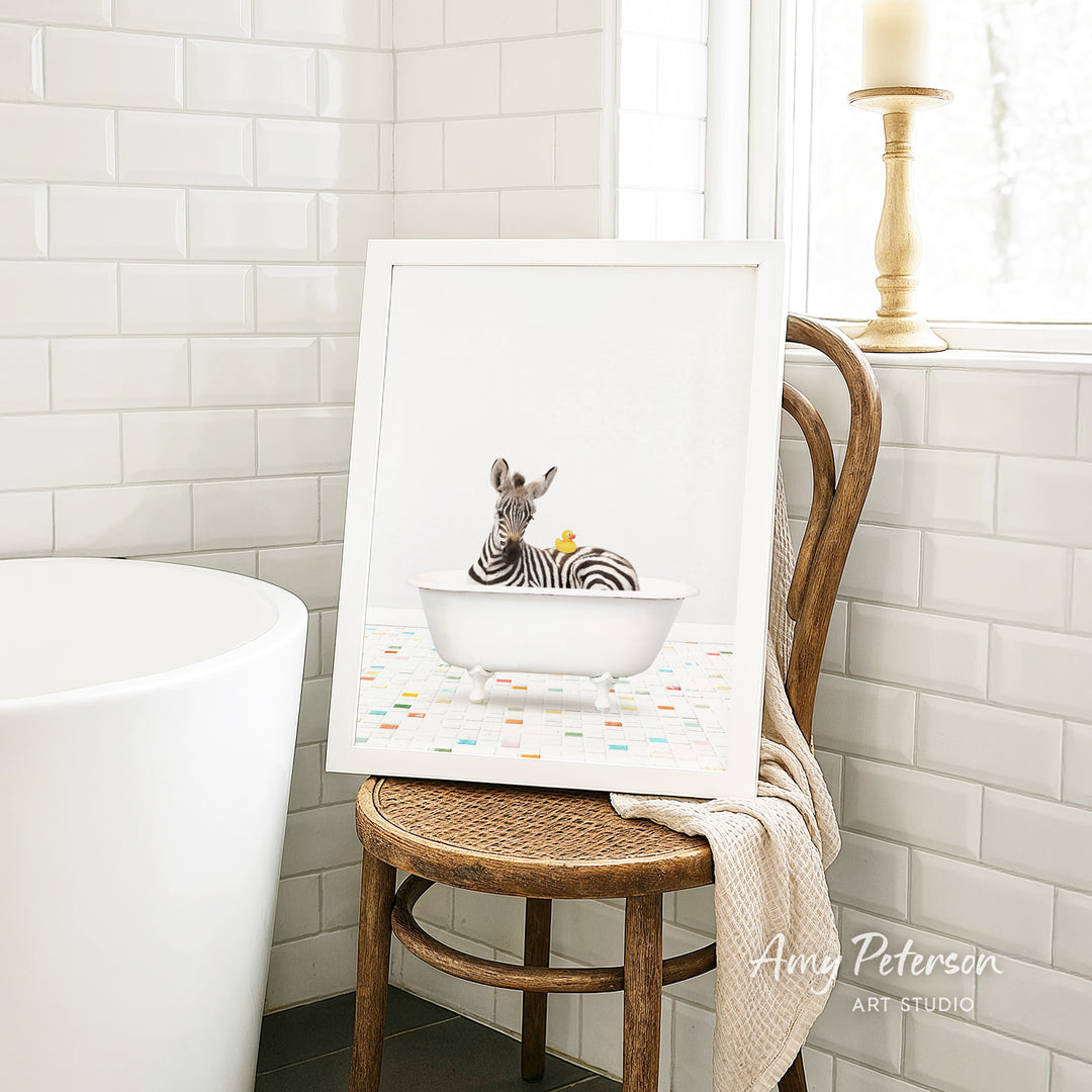 A framed image of a zebra sitting in a white bathtub, with a towel draped over the back of a wooden chair in a bathroom setting.