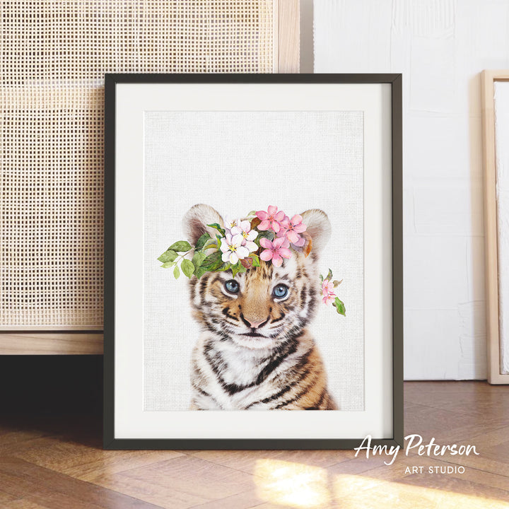A framed print of a tiger cub wearing a floral headpiece is displayed on a wooden floor.