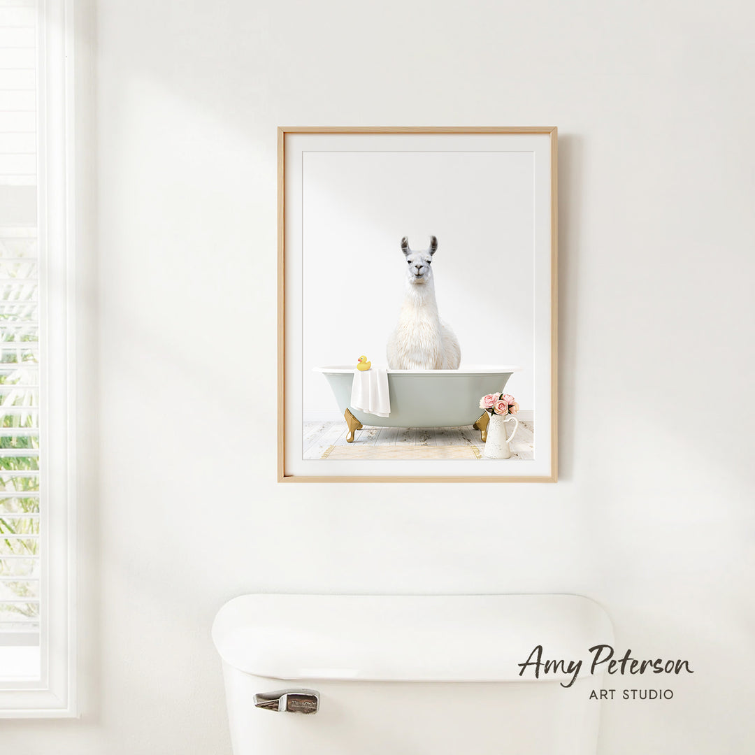 A framed image of a white llama sitting in a bathtub, surrounded by flowers and a vase, is hanging on a white wall in a bathroom.