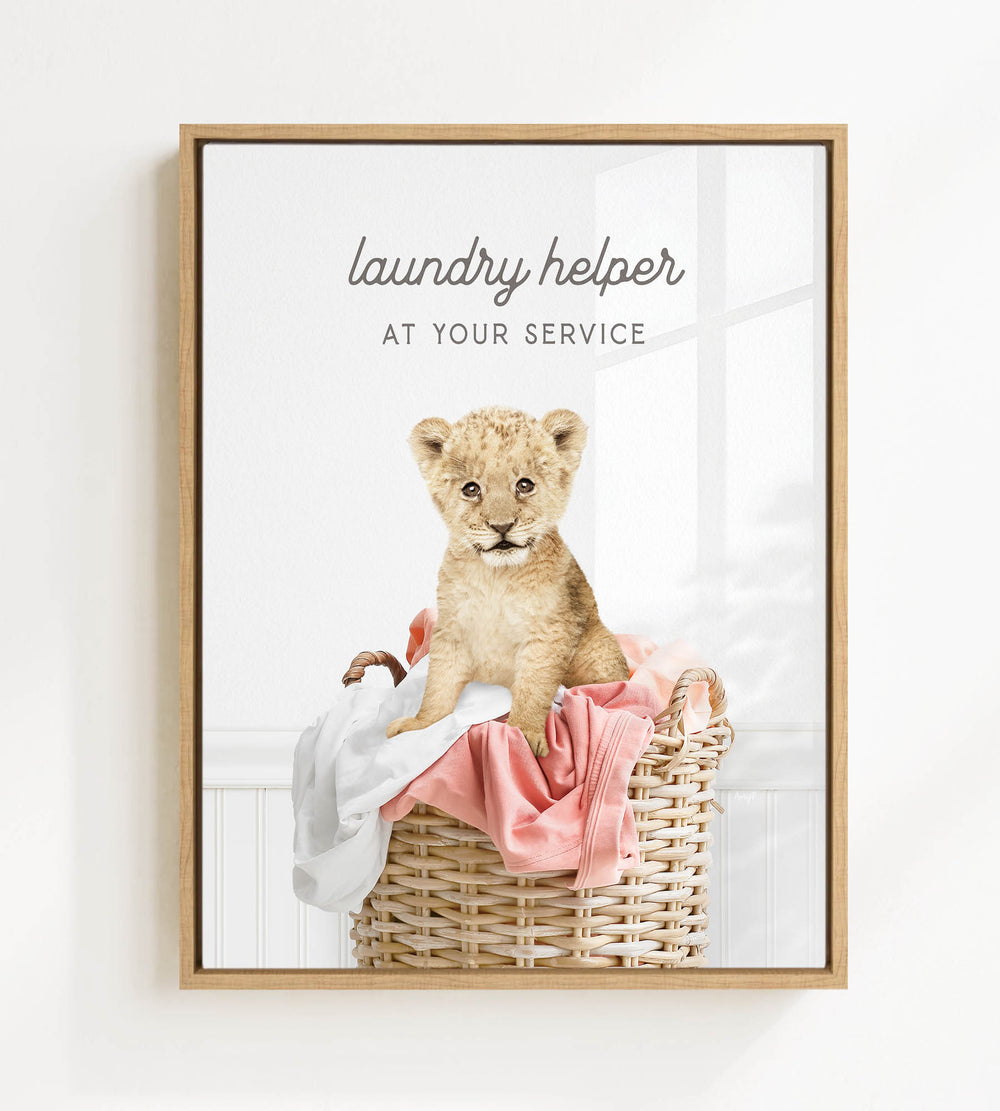 A young lion cub sitting in a wicker laundry basket, with a pink blanket and a sign that says "laundry helper at your service".