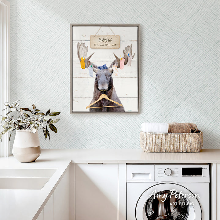 a framed picture of a moose hanging on a wall above a laundry basket, with a potted plant and a white sink in the foreground.
