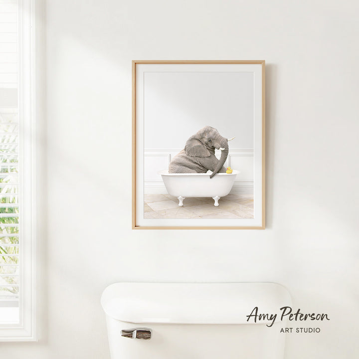 A framed image of a baby elephant sitting in a bathtub, with a window and a toilet visible in the background.