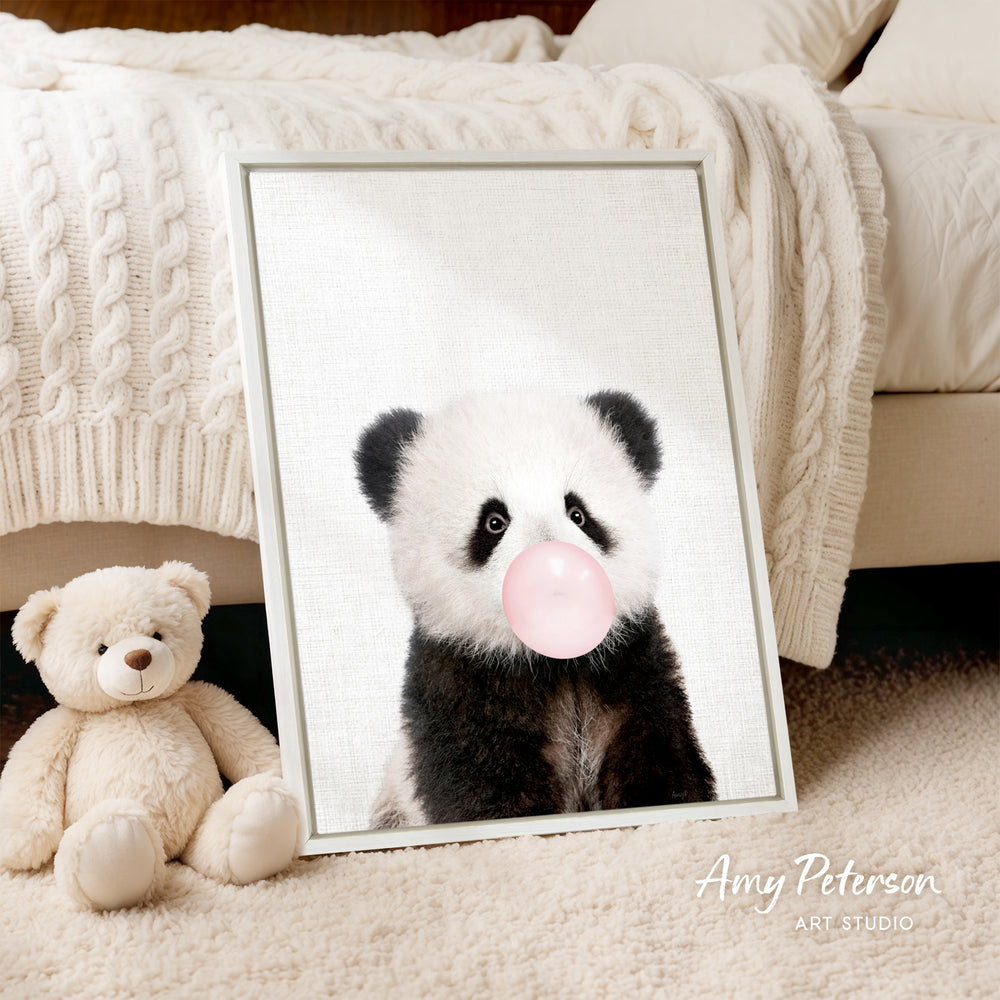 A framed image of a panda blowing a bubble gum bubble is displayed on a bed, accompanied by a teddy bear.