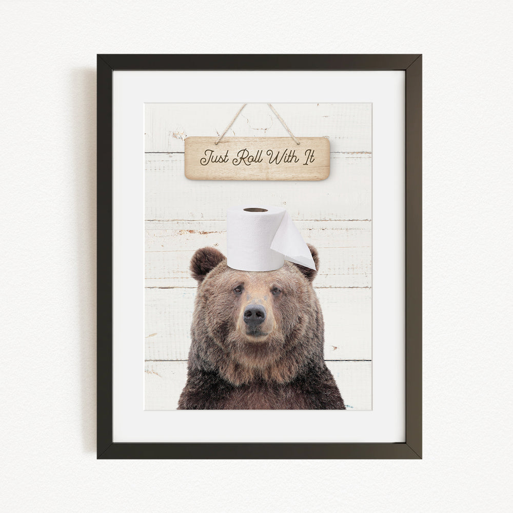 A framed image of a brown bear wearing a toilet paper hat, with a wooden sign above it that says "Just Roll With It".