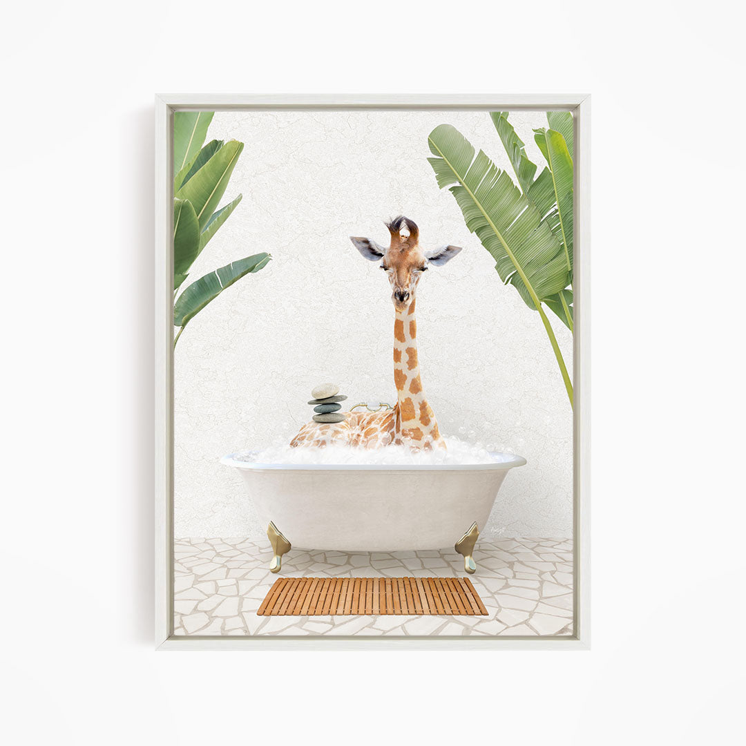A giraffe is sitting in a white bathtub filled with bubbles, surrounded by green leaves and a wooden mat on the floor.