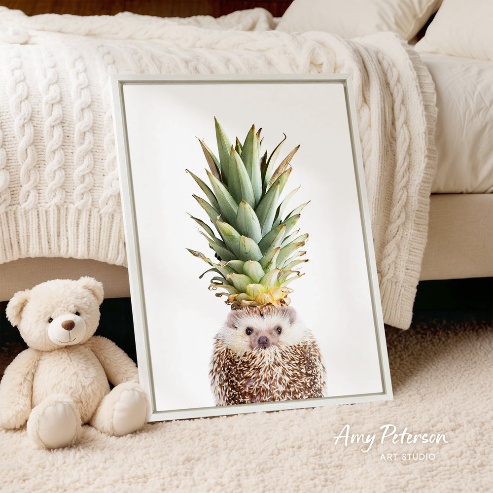 A framed print of a hedgehog wearing a pineapple crown is displayed on a bed, accompanied by a teddy bear.