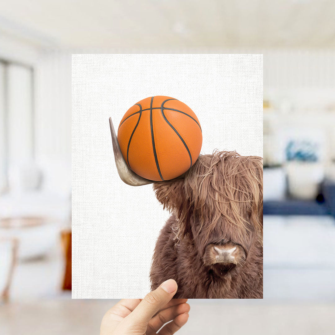 A hand holds up a photograph of a Highland cow with a basketball on its head, creating a playful and whimsical scene.